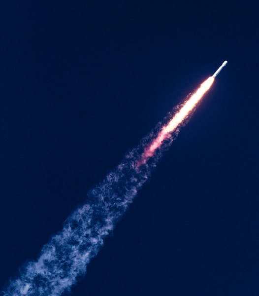 A rocket is ascending into the sky, leaving behind a fiery trail and plume of smoke against a deep blue background.