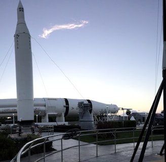 A panoramic view of a rocket garden displaying several space rockets, set in an outdoor museum environment. Tall, white rockets are stationed upright among palm trees and walkways, with a clear sky overhead. Visitors can be seen walking around, and the composition gives a sense of grandeur and human achievement in space exploration.
