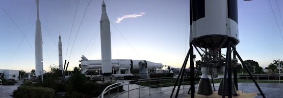 A panoramic view of a rocket garden displaying several space rockets, set in an outdoor museum environment. Tall, white rockets are stationed upright among palm trees and walkways, with a clear sky overhead. Visitors can be seen walking around, and the composition gives a sense of grandeur and human achievement in space exploration.