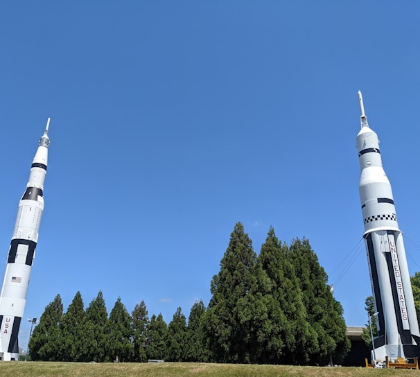Two large rockets stand upright on a grassy area. The rockets are designed with a white and black color scheme and feature the words 'USA' and 'UNITED STATES' on their sides. They are surrounded by a row of tall green trees, under a clear blue sky.