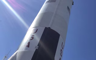 A large rocket standing upright against a clear blue sky. The rocket is painted white with black and red accents, and the word 'USA' is prominently displayed along with a U.S. flag. Sunlight creates a lens flare across the image.