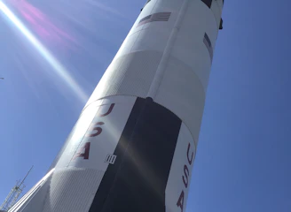 A large rocket standing upright against a clear blue sky. The rocket is painted white with black and red accents, and the word 'USA' is prominently displayed along with a U.S. flag. Sunlight creates a lens flare across the image.
