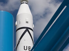 A white rocket with black markings is pointing towards the sky, framed against a backdrop of scattered clouds. In the foreground, there are large blue metallic structures, possibly part of a launch pad or support system.