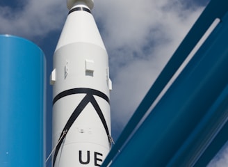 A white rocket with black markings is pointing towards the sky, framed against a backdrop of scattered clouds. In the foreground, there are large blue metallic structures, possibly part of a launch pad or support system.